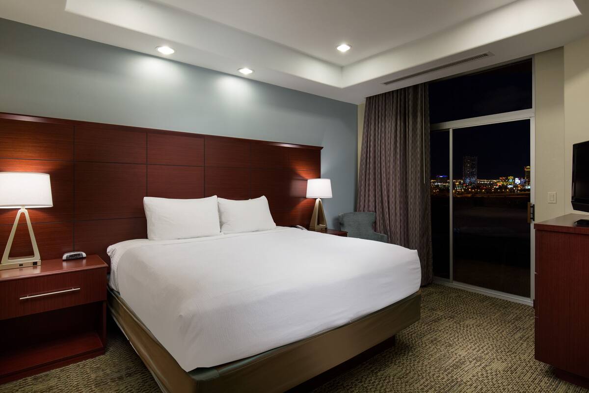 A hotel room with a large bed, a nightstand, a lamp, a chair, a TV, and a view of the city at night.