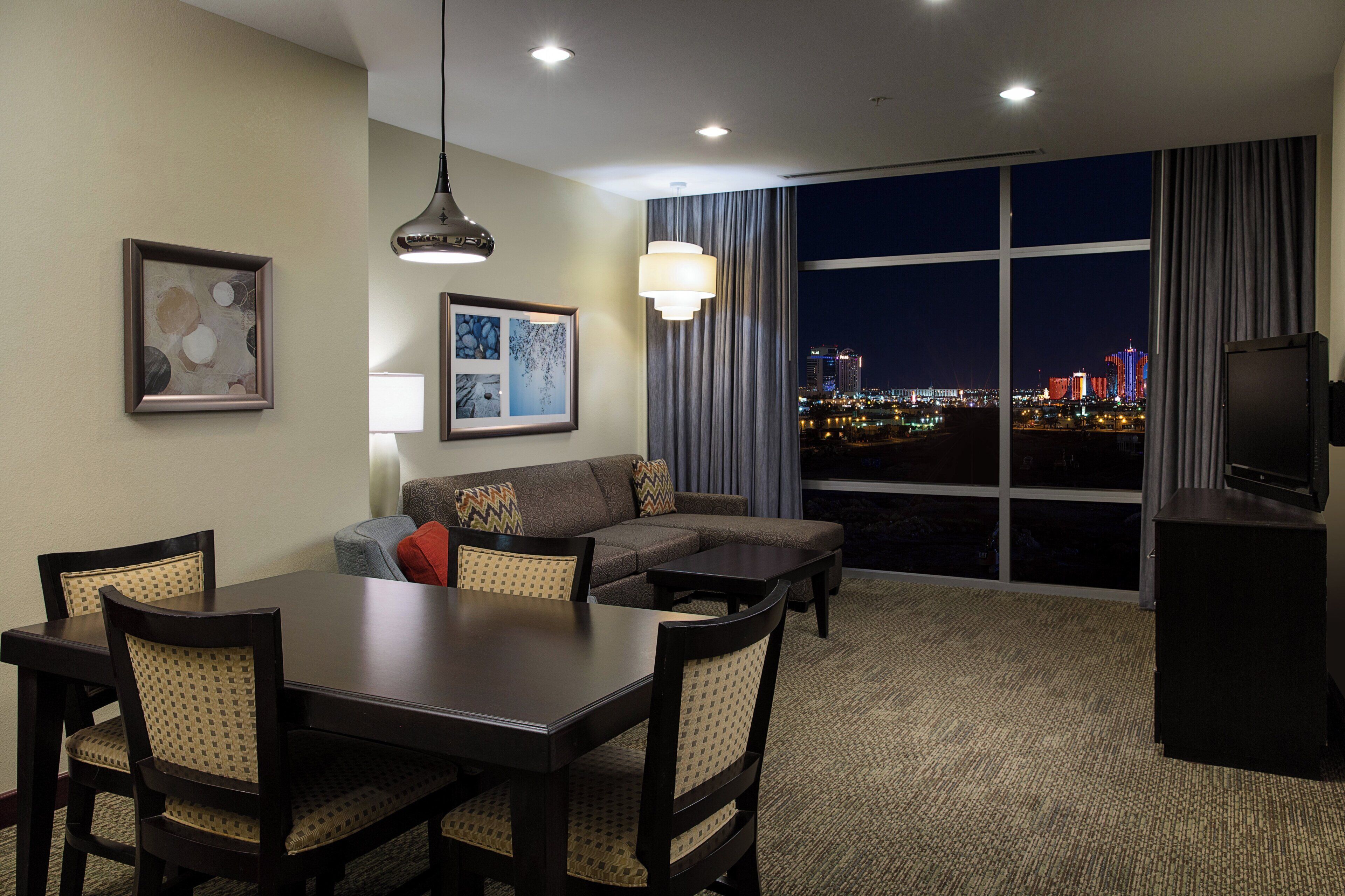 A hotel room with a dining area, a sofa, a TV, and a large window with a city view.