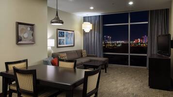 A hotel room with a dining area, a sofa, a TV, and a large window with a city view.