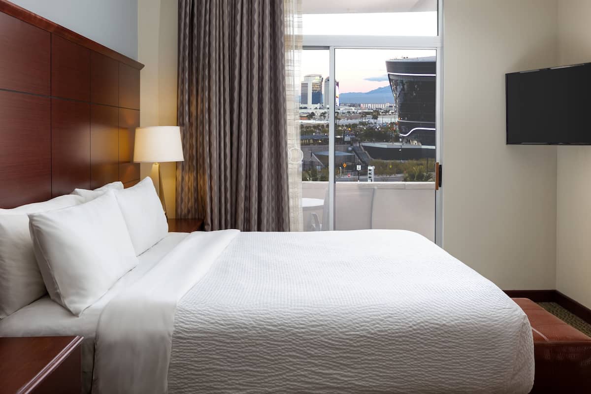 A hotel room with a large bed, a wooden headboard, a window with curtains, and a view of the cityscape.