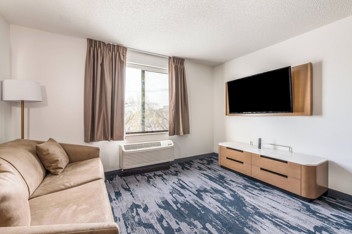 A modern hotel room with a beige sofa, a flat-screen TV mounted on the wall, a wooden sideboard, and a large window with curtains.