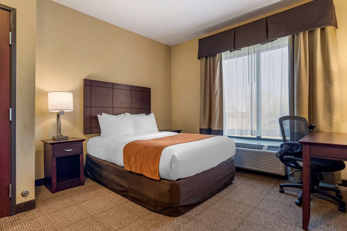Une chambre d’hôtel avec un grand lit, un bureau, une chaise, une table de chevet avec une lampe et une fenêtre avec des rideaux.