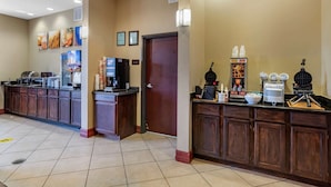 A hotel lobby with a self-service breakfast area, including a coffee machine, waffle maker, and various food items.