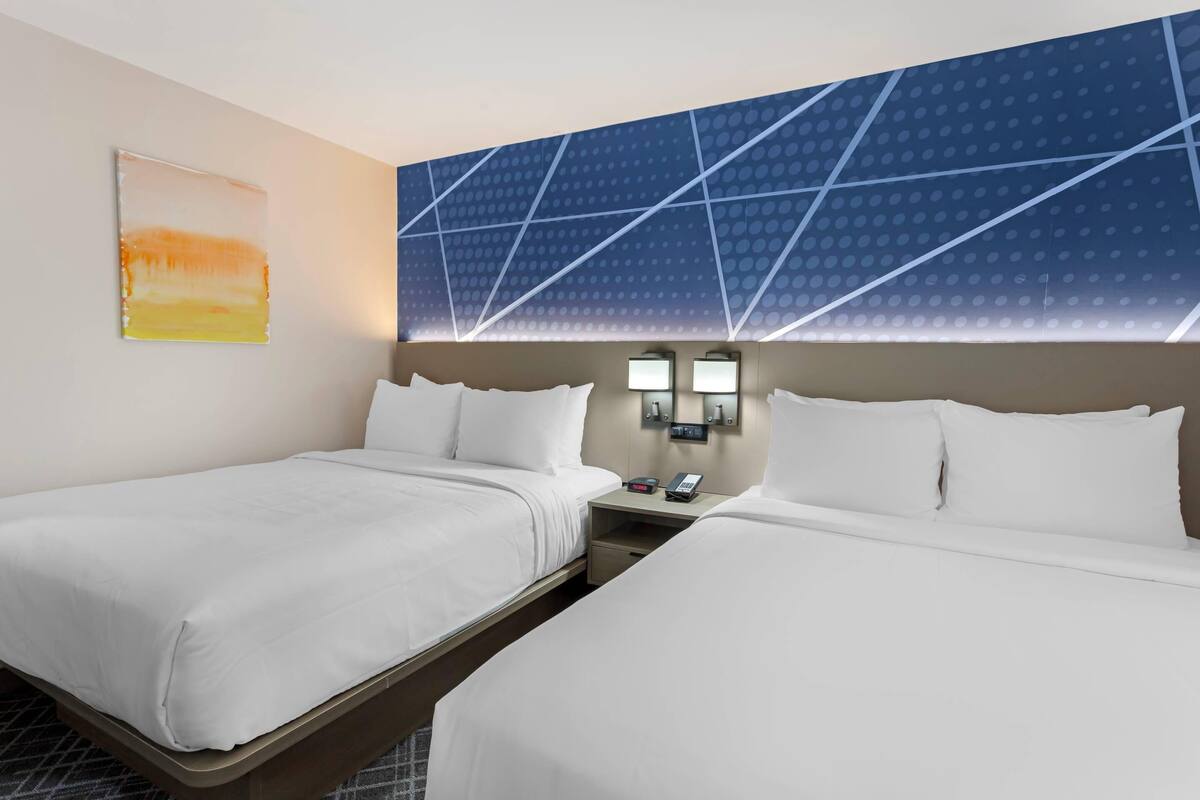A hotel room with two beds, a nightstand with a phone and a lamp, and a wall with a geometric pattern.
