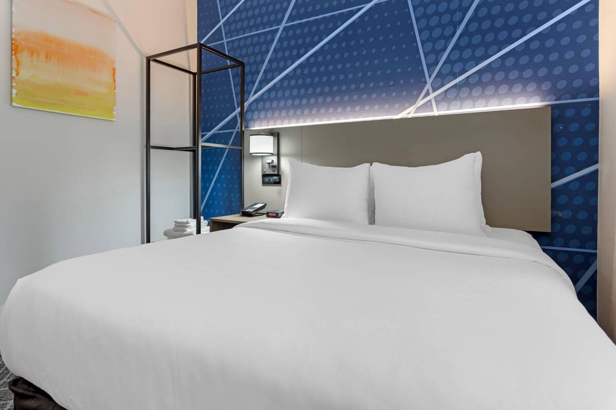 A modern hotel room with a large bed, a nightstand, a lamp, and a wall with a geometric pattern.