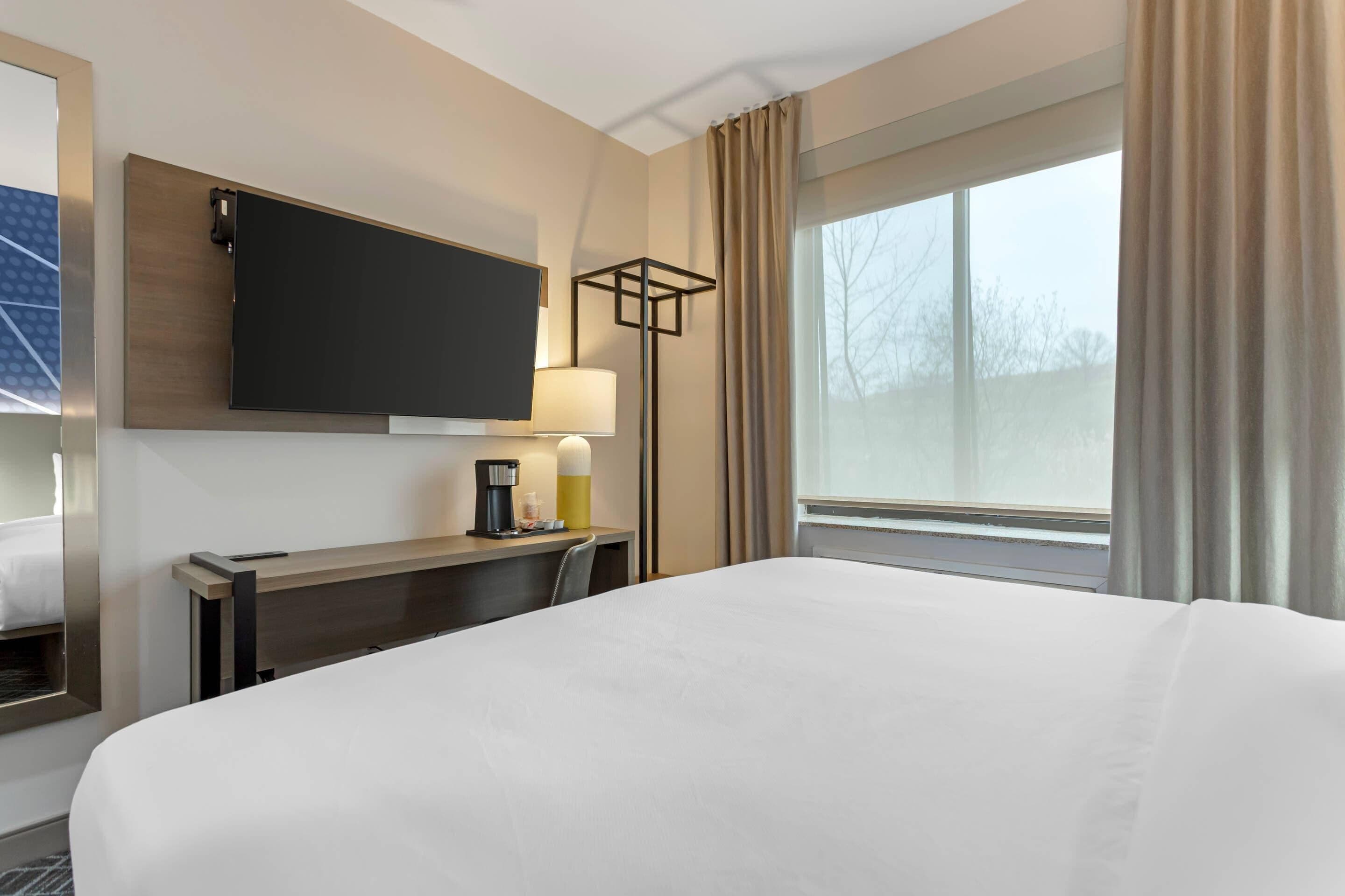 Habitación de hotel con una cama grande, un televisor de pantalla plana en la pared, un escritorio con lámpara y una ventana con cortinas.