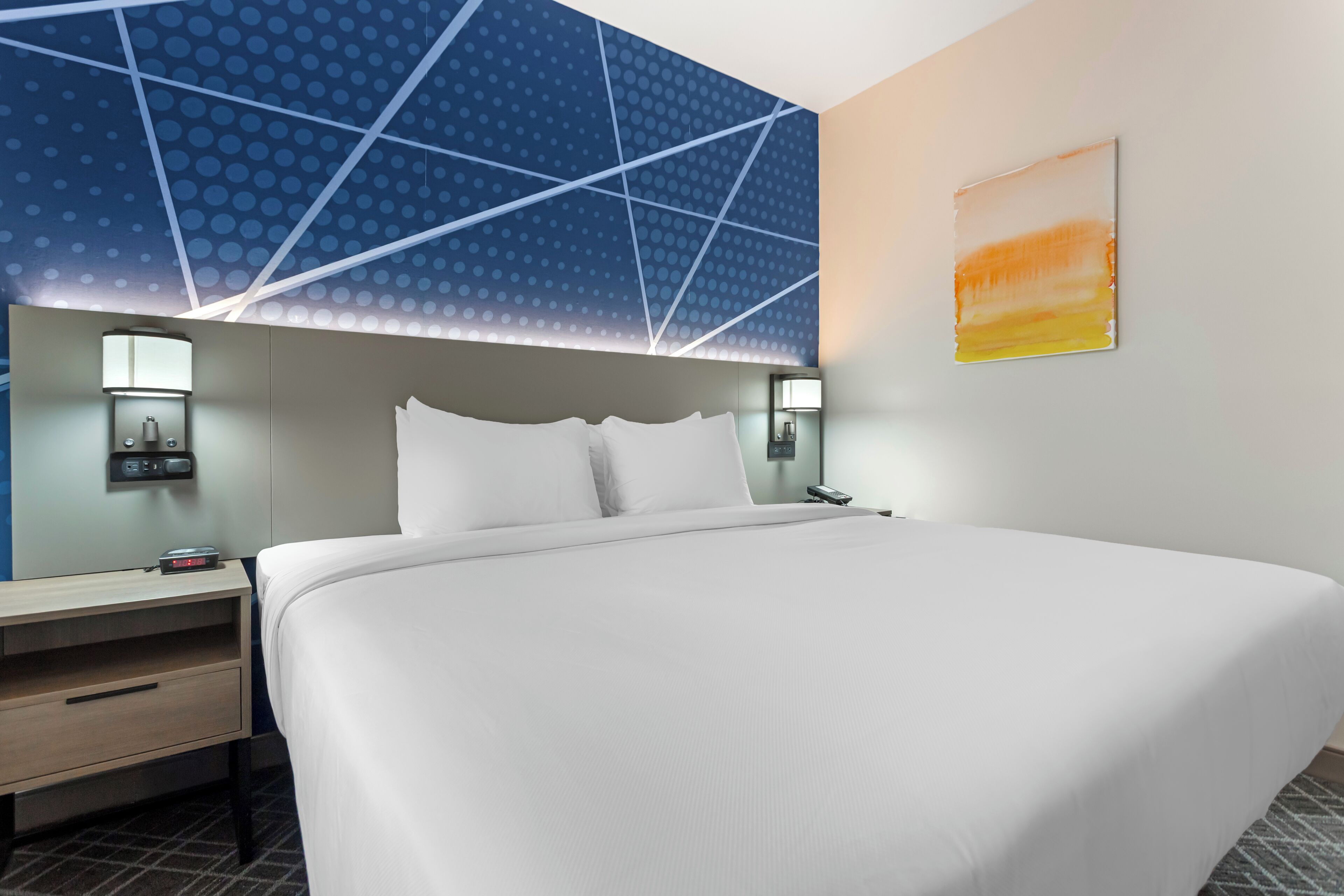 Une chambre d’hôtel moderne avec un grand lit, une table de chevet, une lampe fixée au mur et un tableau.