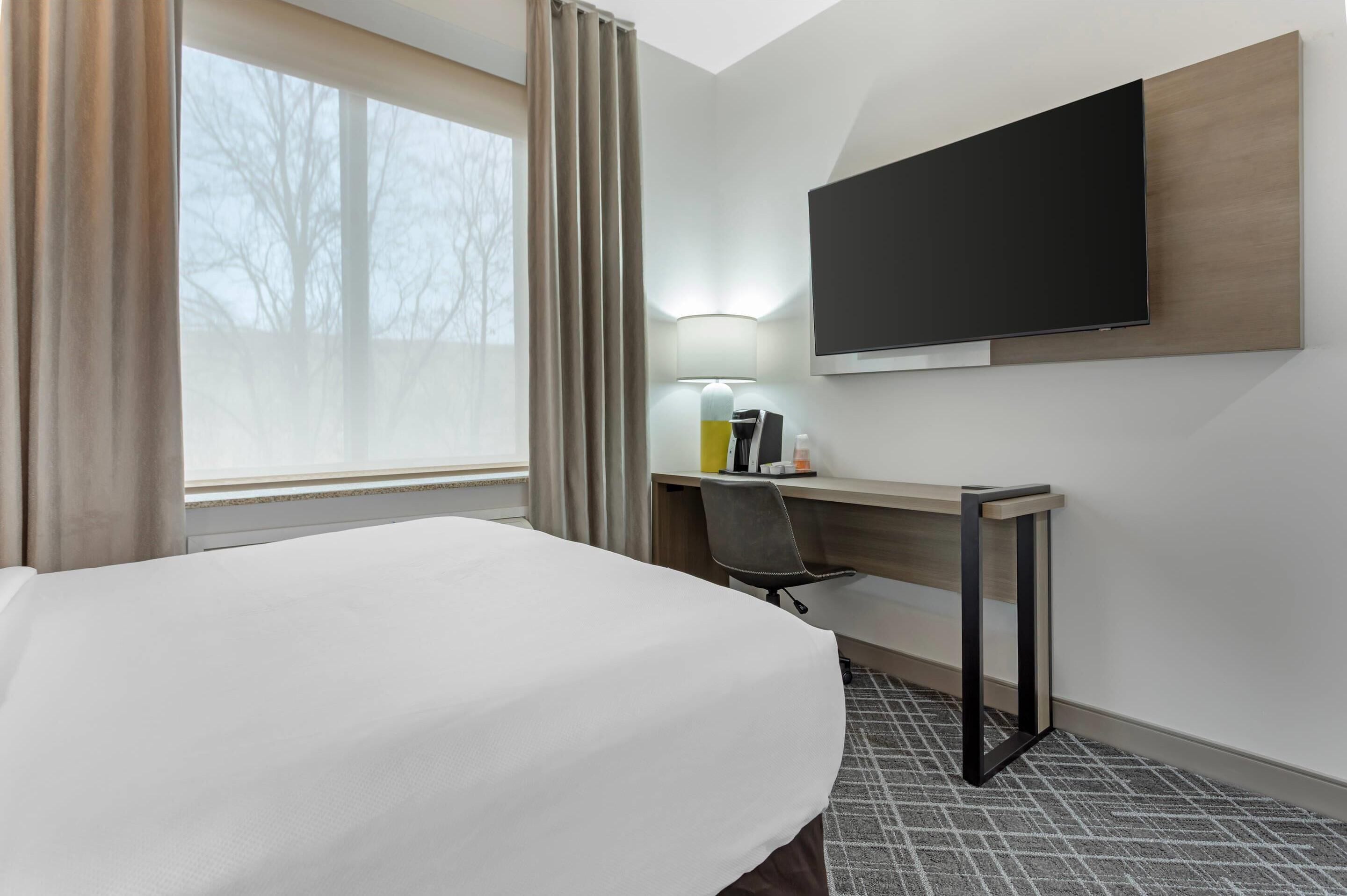 A hotel room with a large bed, a desk with a chair, a flat-screen TV mounted on the wall, and a window with curtains.