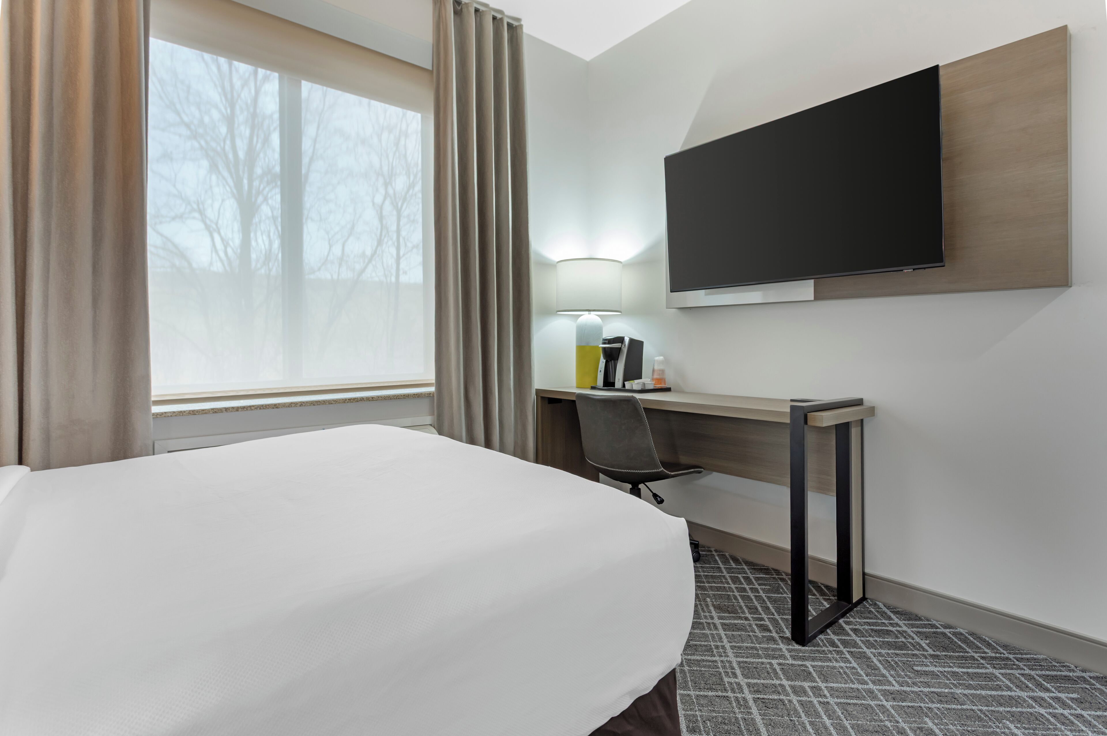 Quarto de hotel com uma cama, escrivaninha, cadeira, televisão na parede e janela com cortinas.