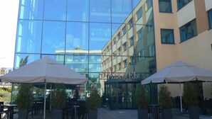A modern hotel with a glass facade, outdoor seating with umbrellas, and a clear view of the surrounding cityscape. - Best Western Premier Hotel Regensburg (Regensburg)