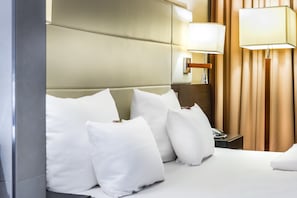 A hotel room with a bed, white pillows, a wooden headboard, a bedside table with a lamp, and a telephone.