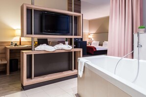 A modern hotel room with a TV mounted on a wooden console, a bathtub, a desk with a lamp, and a bed with a red blanket.
