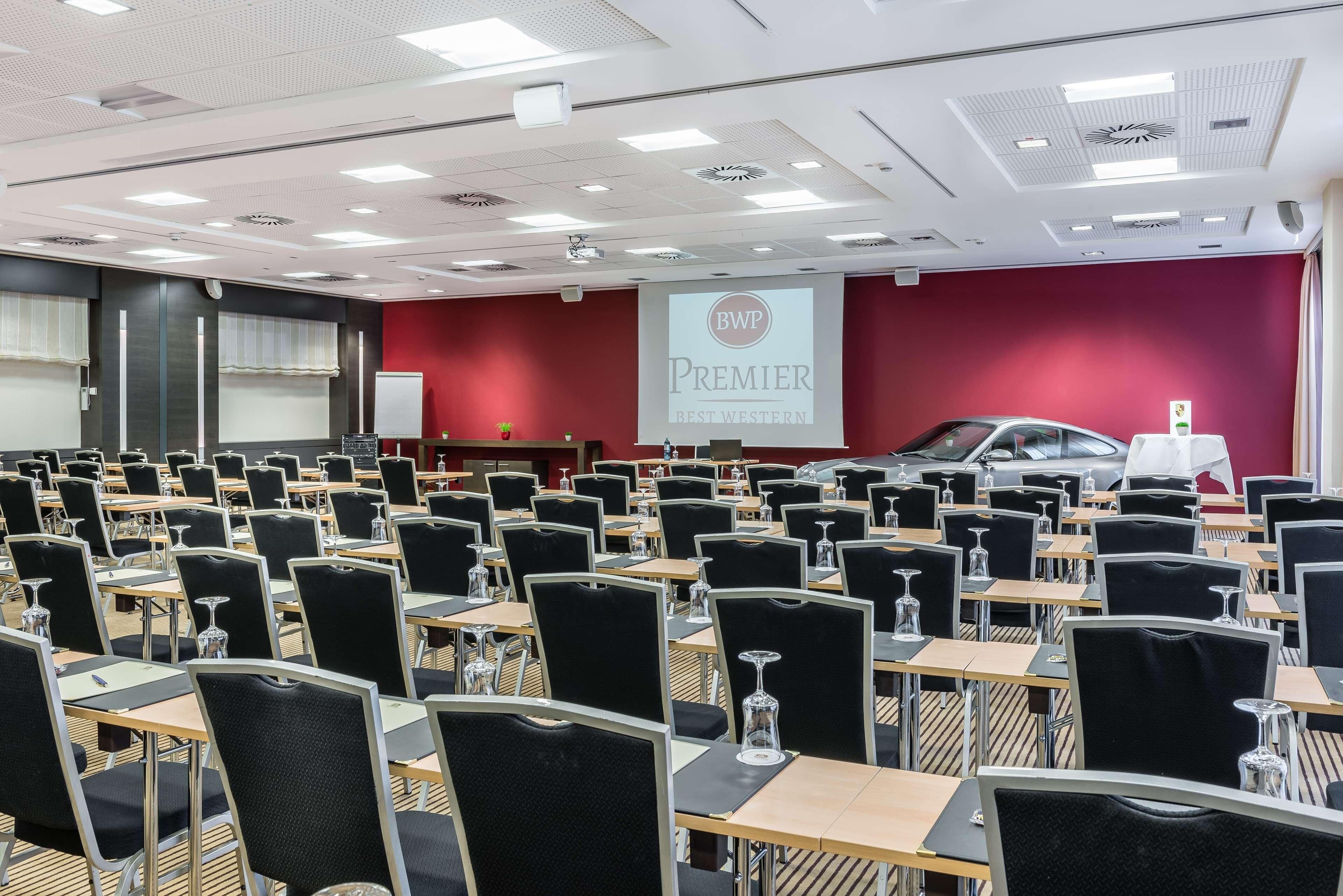 A conference room with rows of tables and chairs, a projector screen, and a red wall with a logo.
