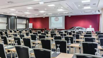 A conference room with rows of tables and chairs, a projector screen, and a red wall with a logo.