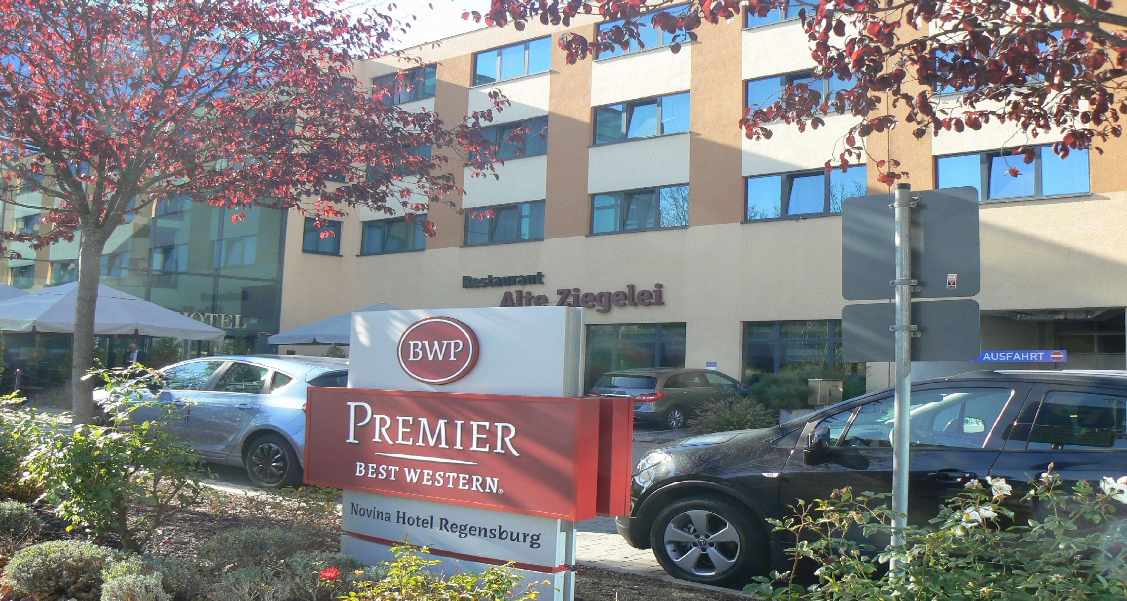Ein Hotelschild mit der Aufschrift 'Premier Best Western' und 'Novina Hotel Regensburg', ein Gebäude mit dem Schild 'Restaurant Alte Ziegleri' sowie abgestellte Autos.