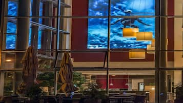 An outdoor seating area with tables and chairs, a large digital screen displaying a sky and clouds, and hanging lamps.