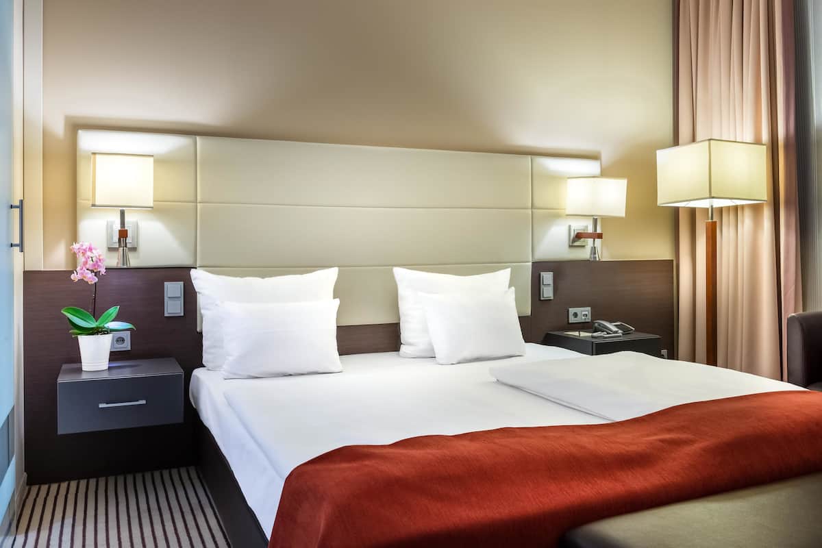 Une chambre d’hôtel avec un grand lit, deux tables de chevet, une petite table, une lampe et une plante en pot.