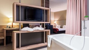 A modern hotel room with a flat-screen TV mounted on a wooden console, a large white bathtub, and a neatly made bed with a red blanket. - Best Western Premier Hotel Regensburg (Regensburg)