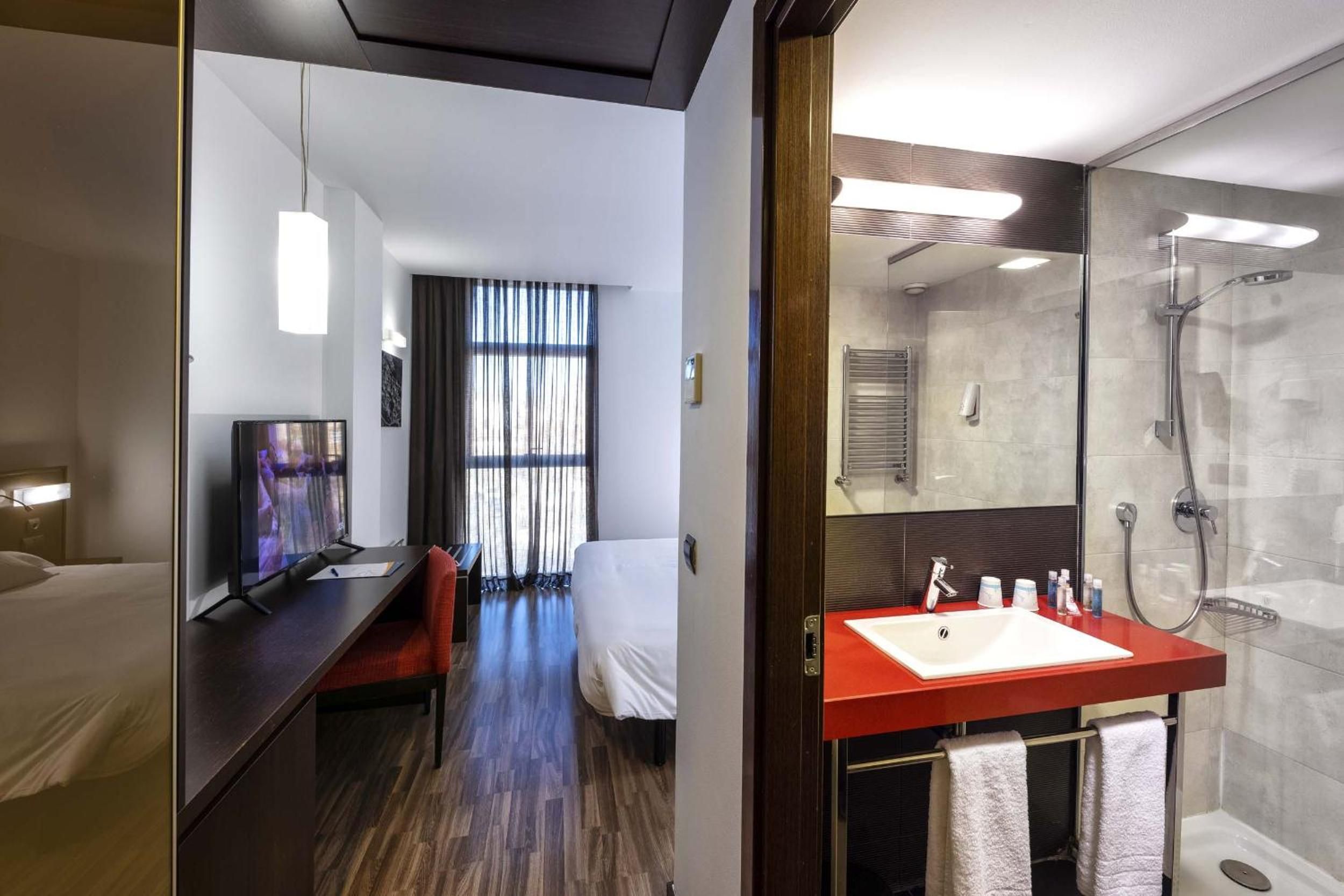 A hotel room with a bed, desk, TV, and a bathroom with a shower and sink.