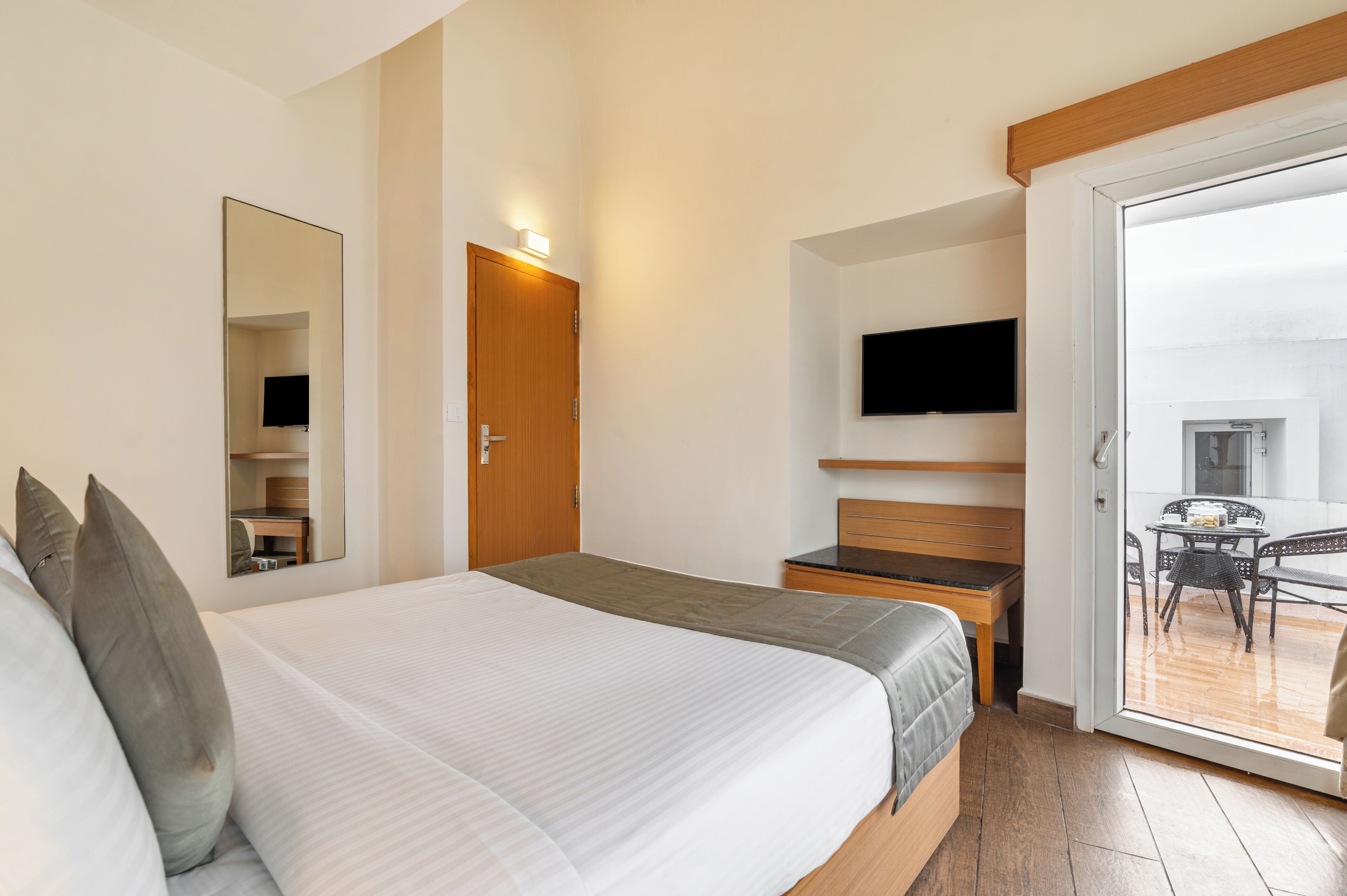 Ein Hotelzimmer mit einem großen Bett, einem Nachttisch aus Holz, einem an der Wand befestigten Fernseher und einer Schiebetür zu einem Balkon mit Tisch und Stühlen.