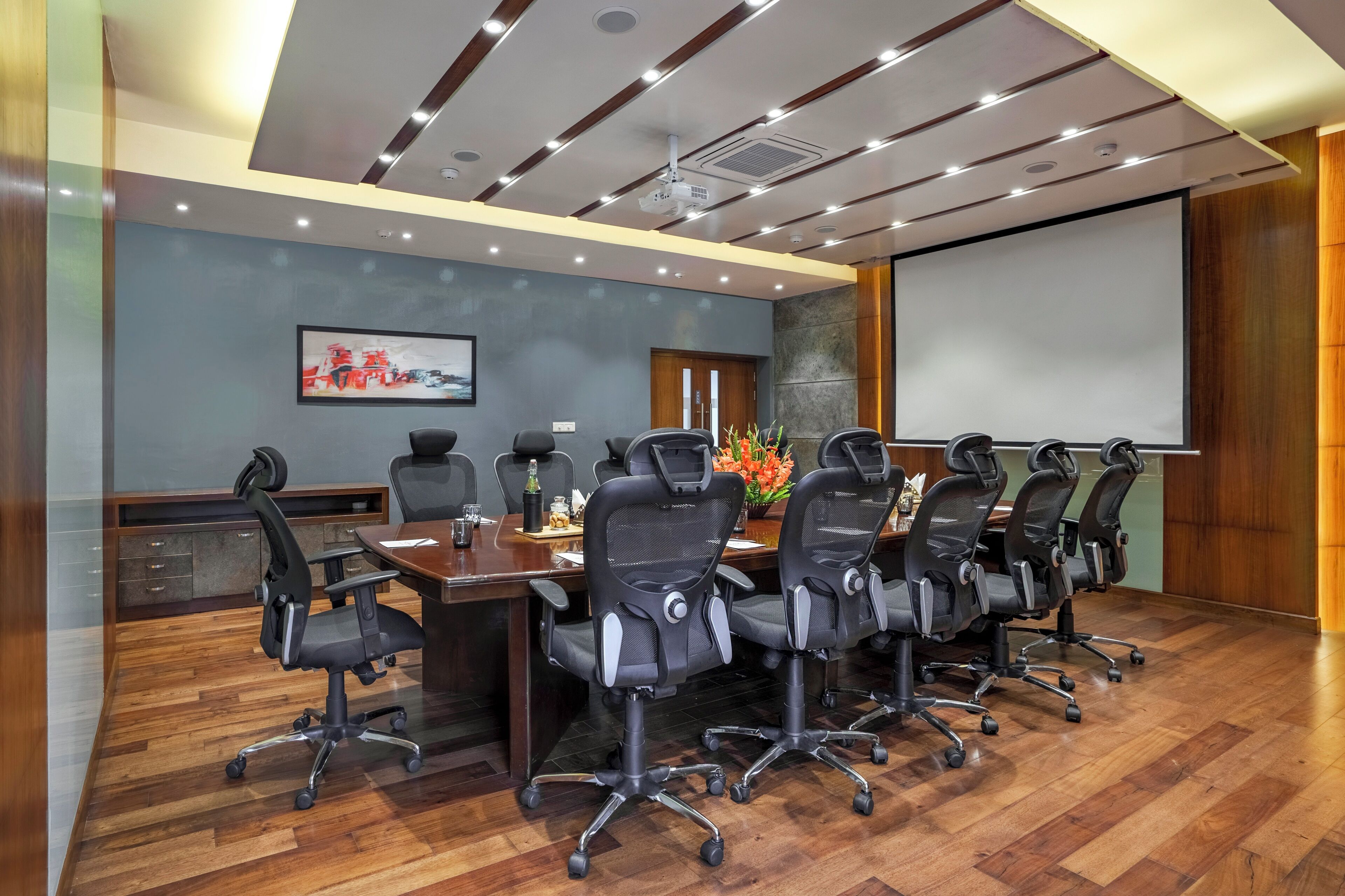 A conference room with a long table, multiple chairs, a projector screen, and a wooden floor.