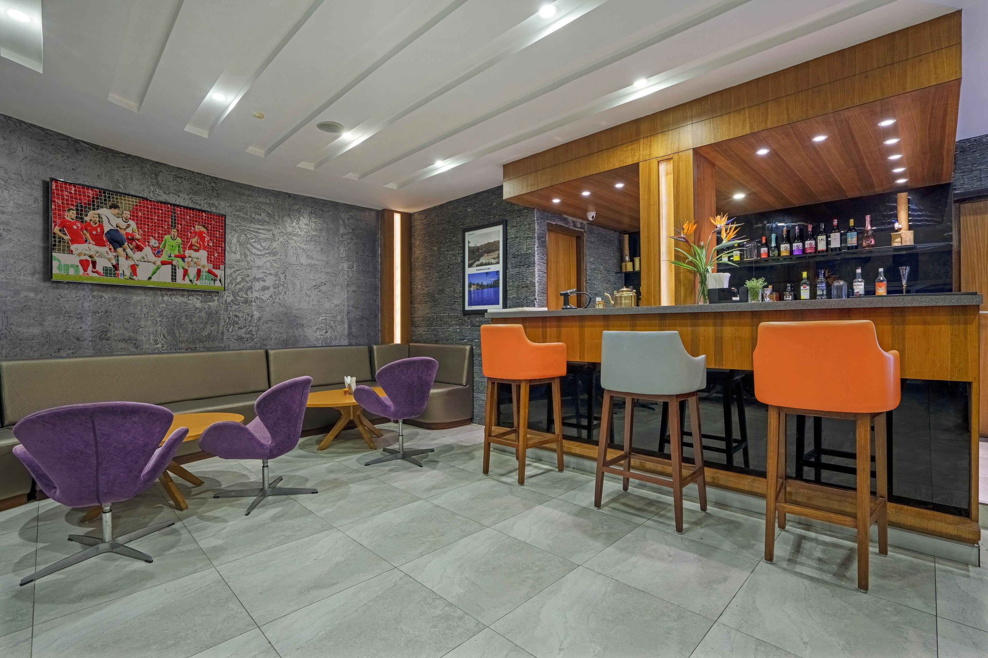 Zona bar moderna con bancone in legno e sgabelli, area salotto con sedie viola e arancioni e decorazioni murali a tema sportivo.