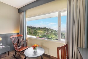 A room with a large window, a wooden rocking chair, a small round table with a vase of flowers, and a view of a green landscape.