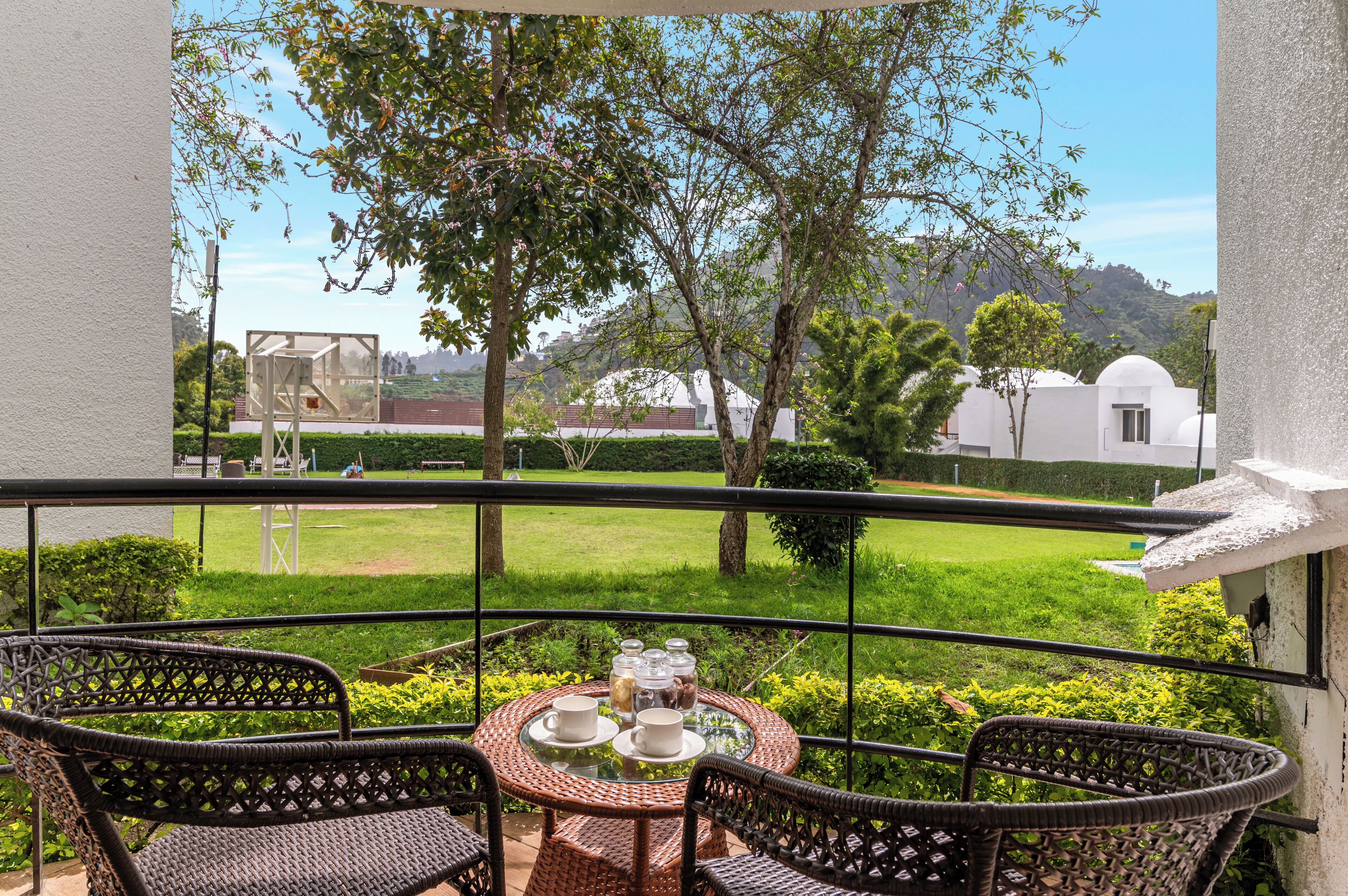 Un balcón con sillas de mimbre y una mesa dispuesta para dos, con vistas a un jardín cuidado y un edificio blanco con cúpula.