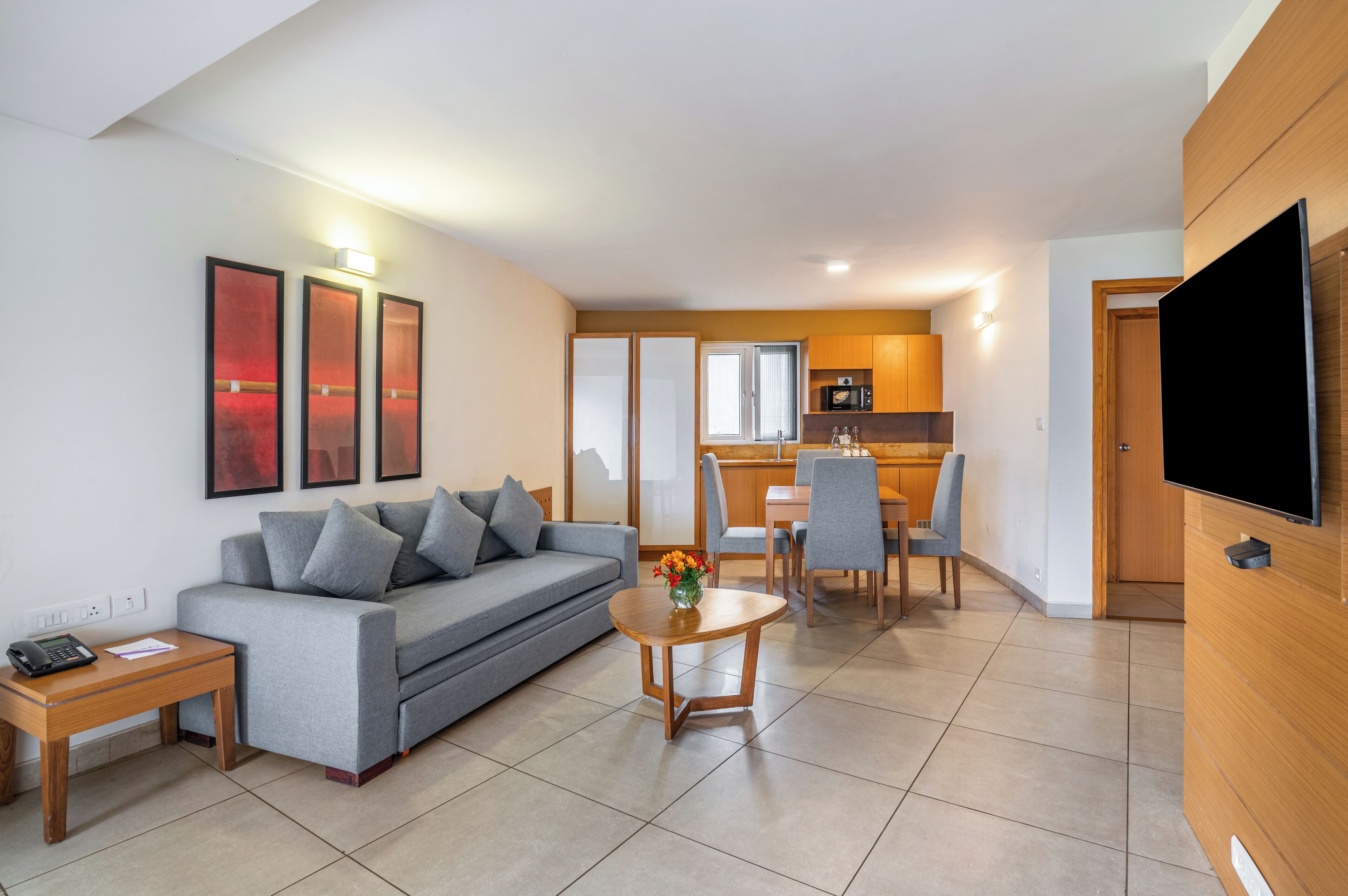 A modern living room with a grey sofa, a wooden coffee table, and a TV on the right. There is a kitchen area with wooden cabinets and a dining table with chairs.