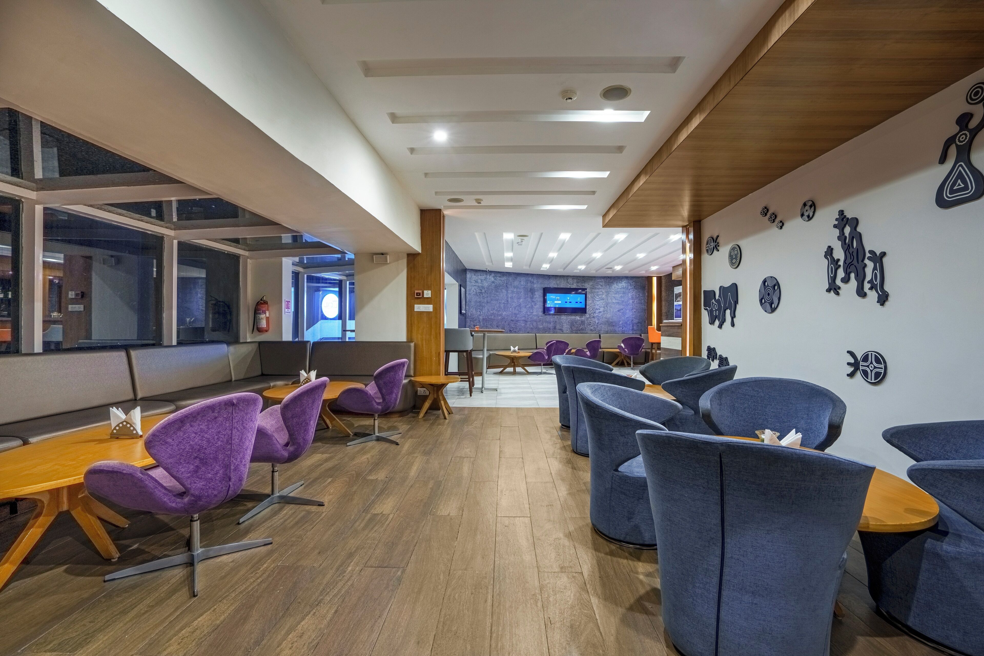 Un hall d’hôtel moderne avec des sièges violets et bleus, un comptoir d’accueil en bois et un téléviseur fixé au mur.