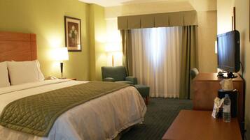 A hotel room with a bed, two chairs, a desk with a television, and a window with curtains.