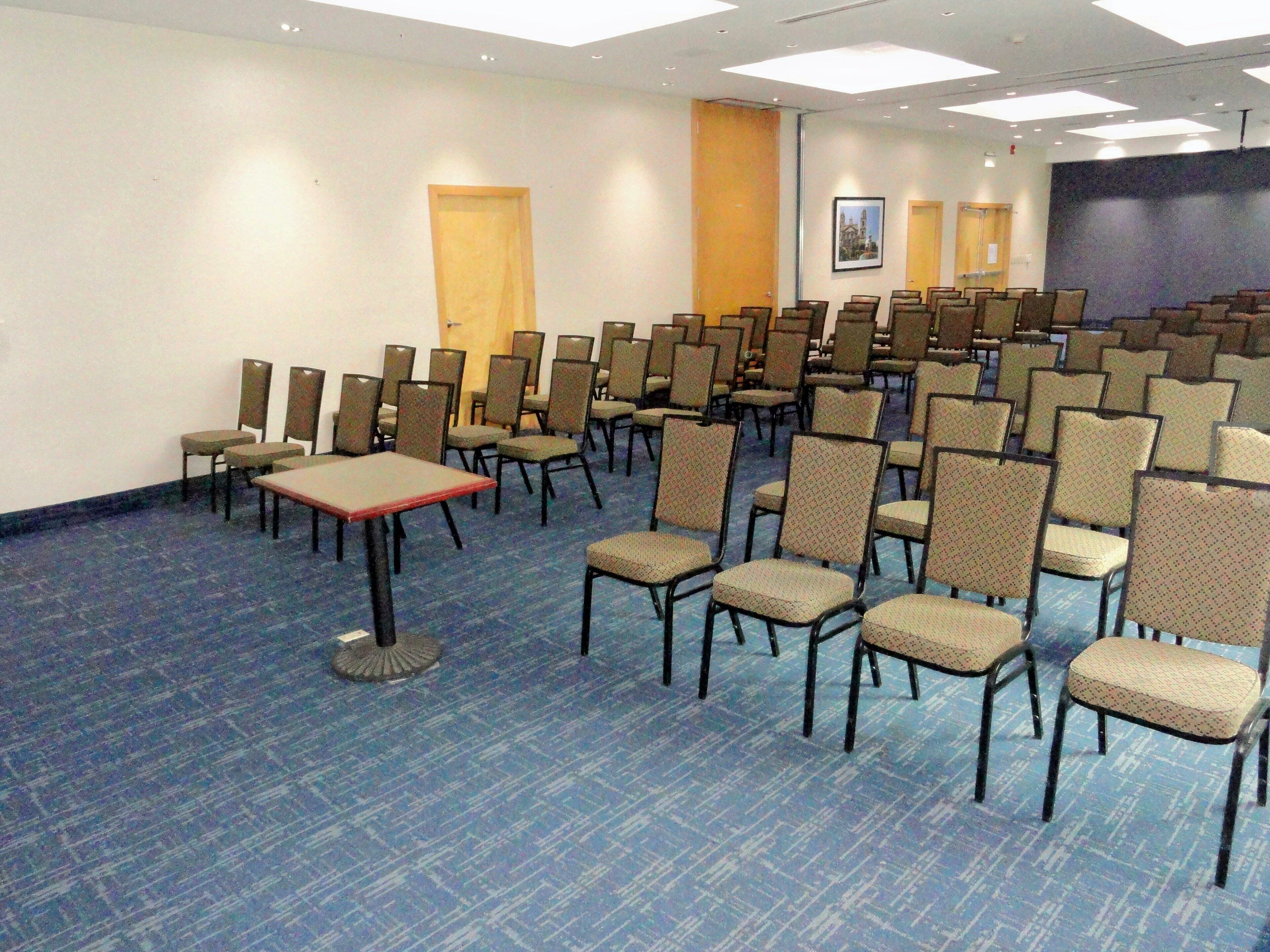 A conference room with rows of chairs and tables, a projector screen, and a door in the background.