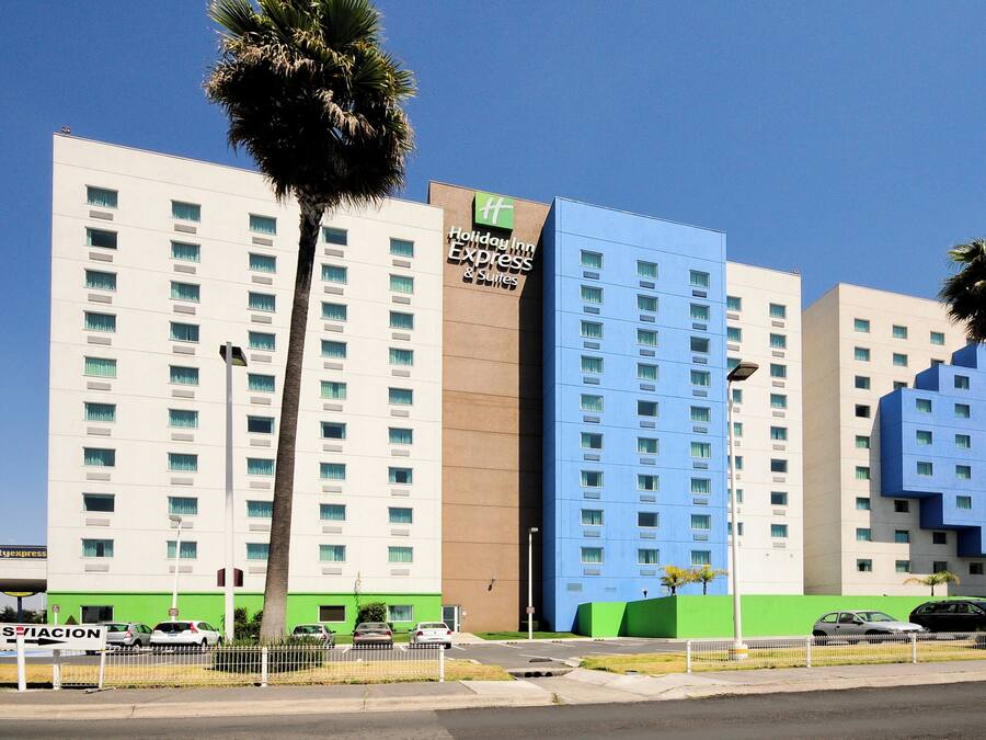 A multi-story hotel with a Holiday Inn Express & Suites sign, featuring a modern design with a mix of white and blue facades, and palm trees in front.