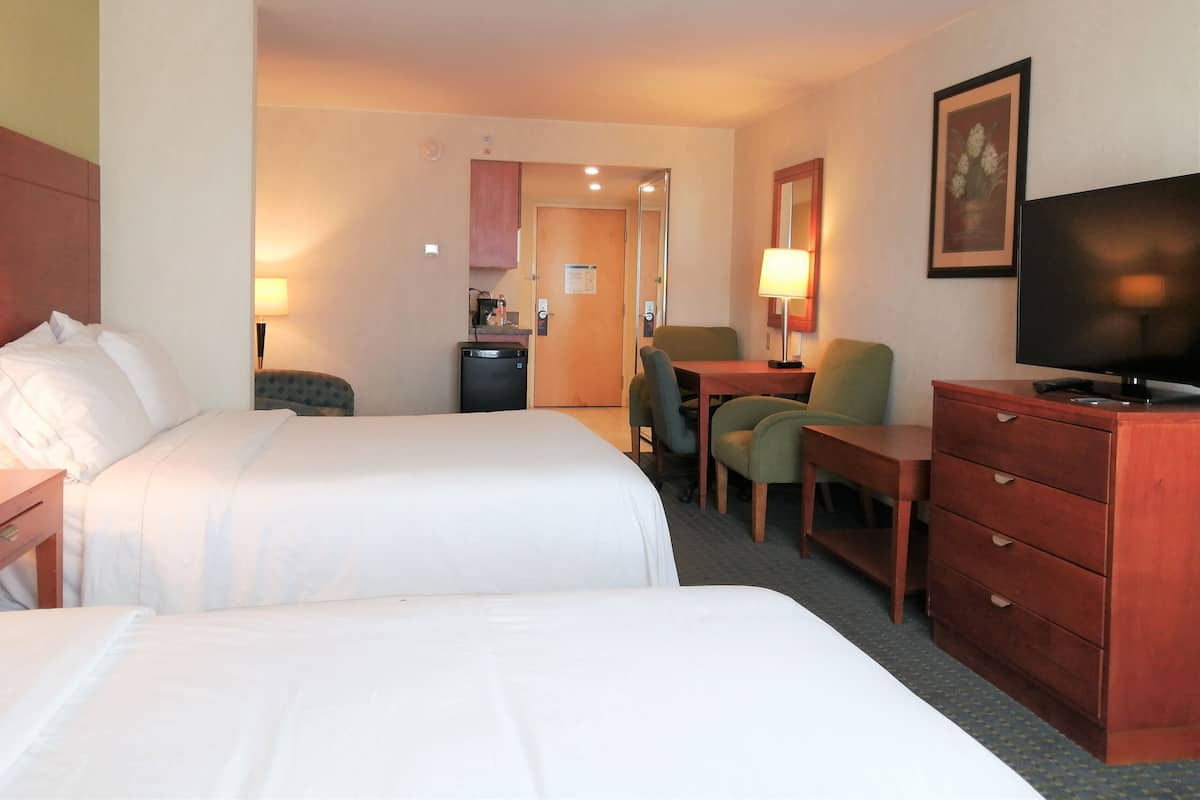 A hotel room with two beds, a desk, a chair, a television, and a small kitchenette.