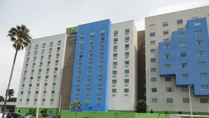 Een meerhoog hotel met een opvallende blauw-beige gevel, met een bord voor 'Holiday Inn Express & Suites' en een bewegwijzer voor 'Salida Blvd. Aeropuerto'.