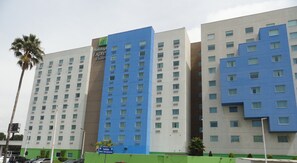 Un hôtel à plusieurs étages, doté d’une façade bleue et beige remarquable, avec une enseigne indiquant « Holiday Inn Express & Suites » et un panneau de signalisation indiquant la direction de « Salida Blvd. Aeropuerto ».