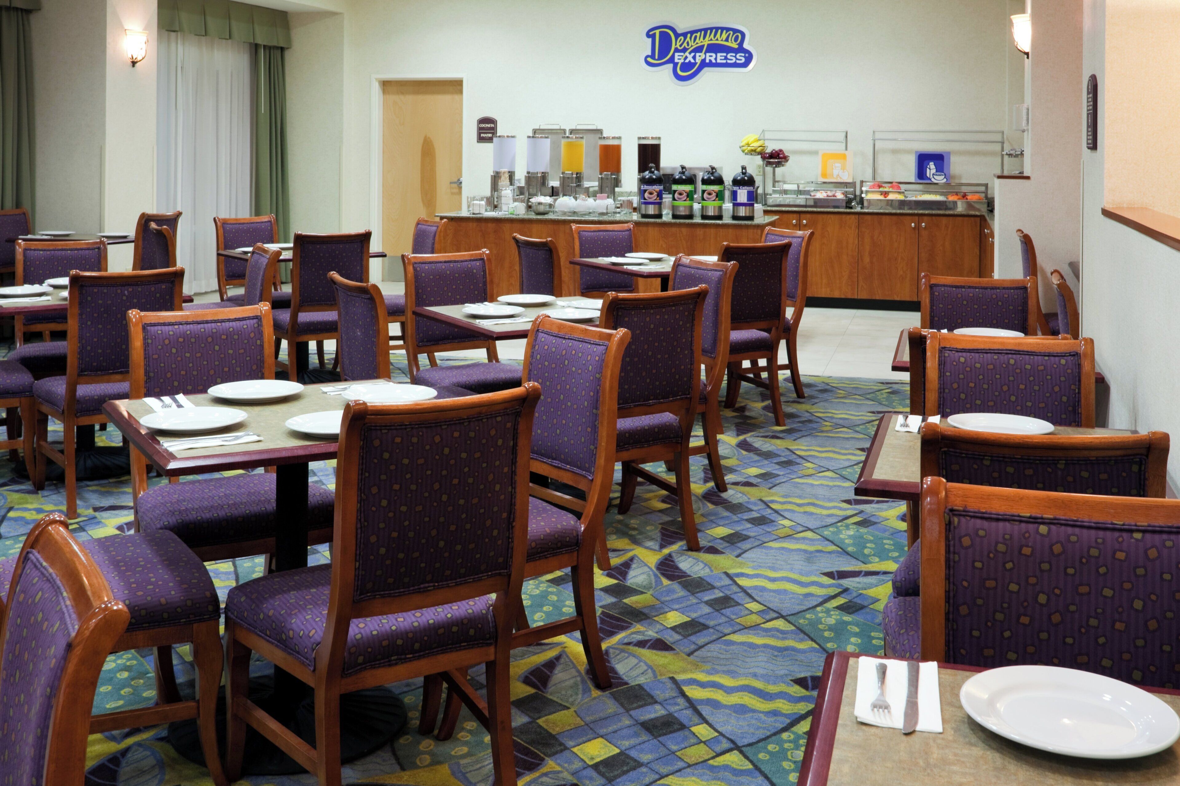 A hotel dining area with purple upholstered chairs and tables, a buffet counter with various food items, and a sign for Desayuno Express.