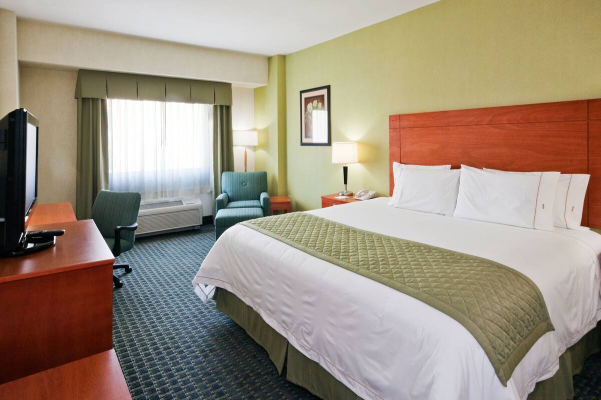A hotel room with a large bed, a desk with a computer, a chair, a TV, and a window with curtains.
