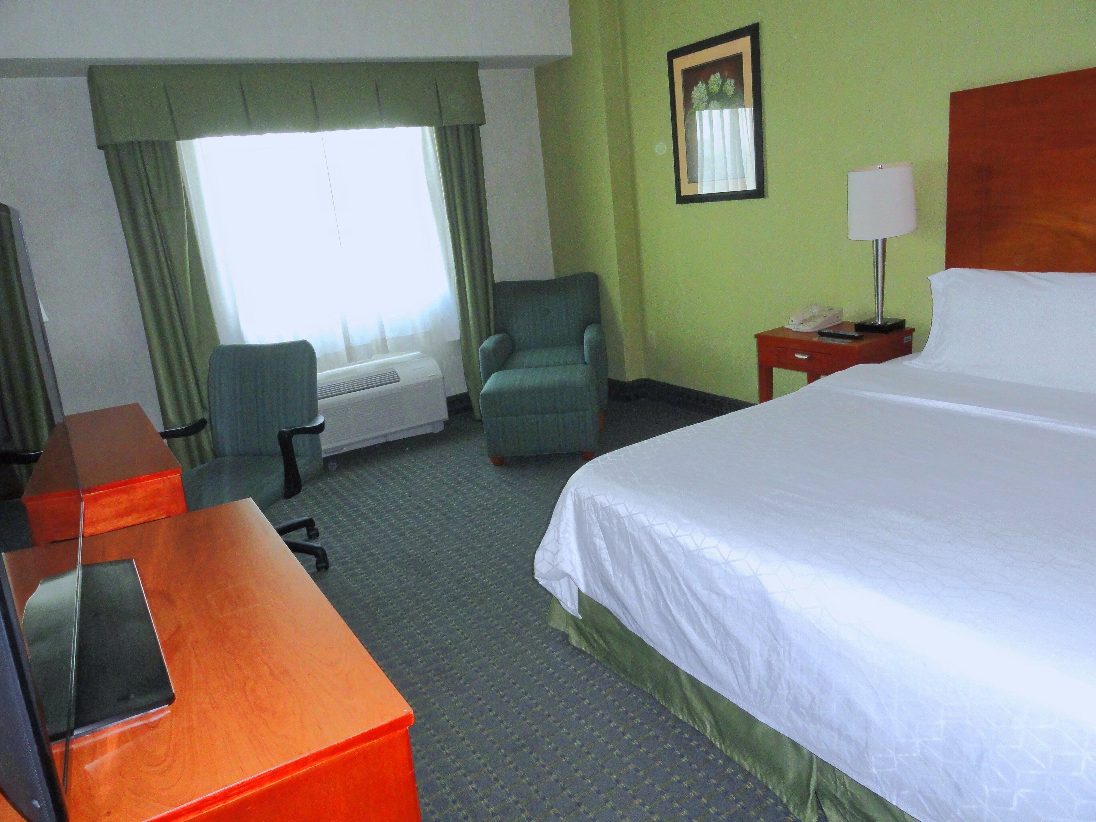 A hotel room with a bed, a desk, a chair, a lamp, and a window with curtains.