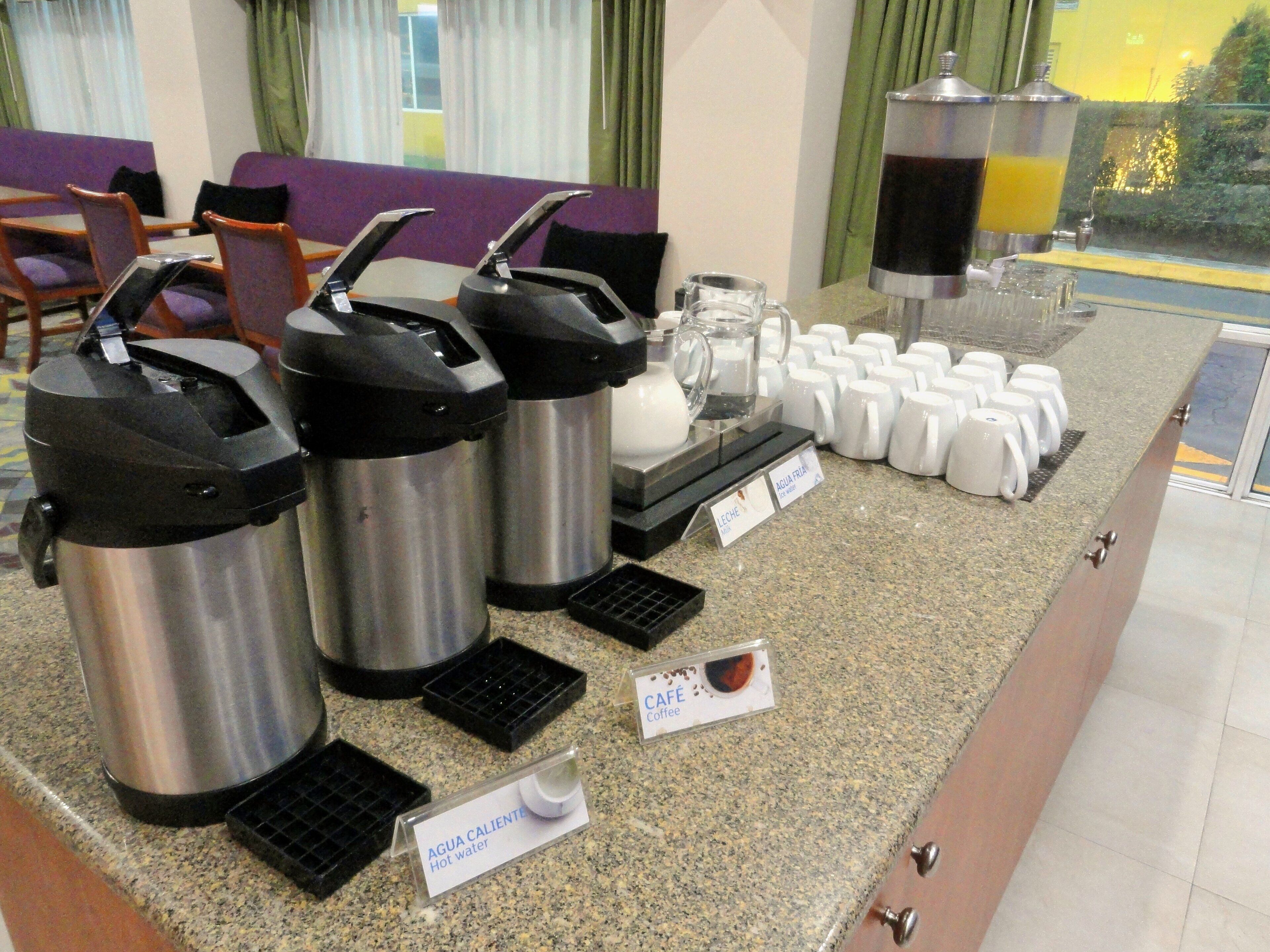 Una estación de bebidas de autoservicio con dispensadores de agua caliente y café, una cafetera y una jarra grande de jugo de naranja.