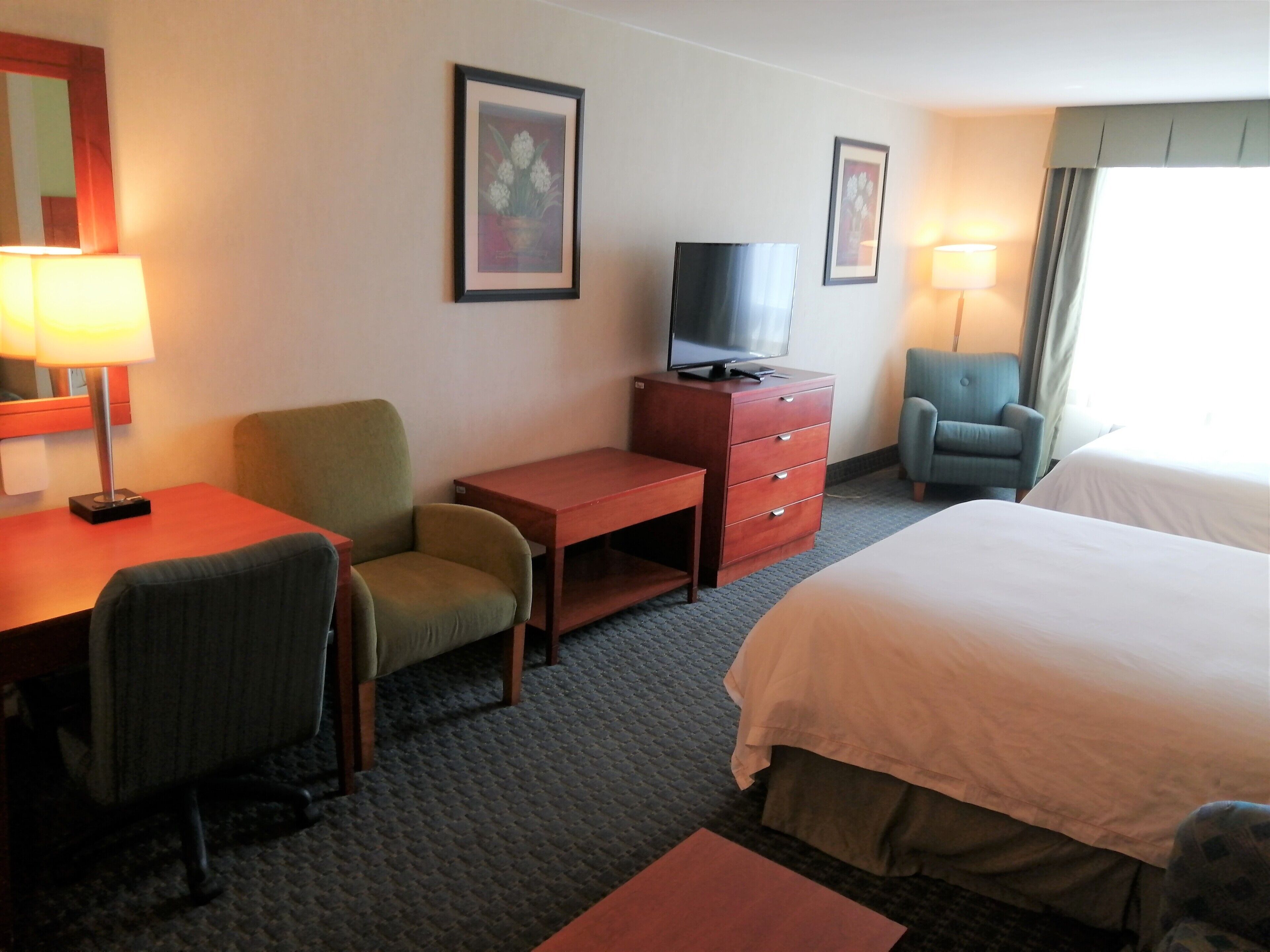 Habitación de hotel con cama, escritorio, silla, cómoda, televisor y dos cuadros en la pared.