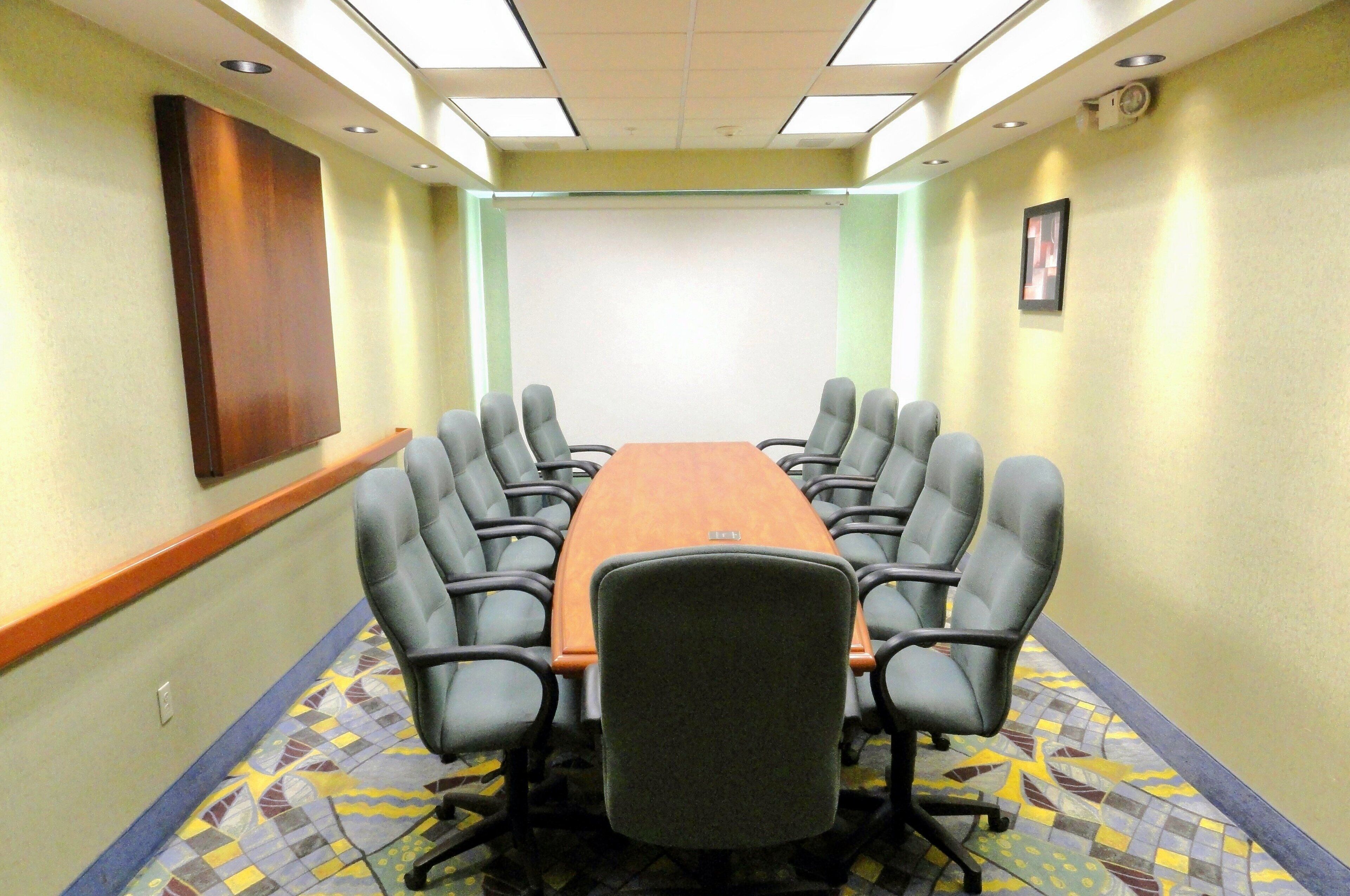 Une salle de conférence équipée d’une longue table et de six chaises, d’un écran de projection et d’un tapis à motifs.