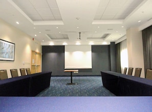Un salón de conferencias con una larga mesa, sillas, una pantalla de proyector e iluminación empotrada en el techo.