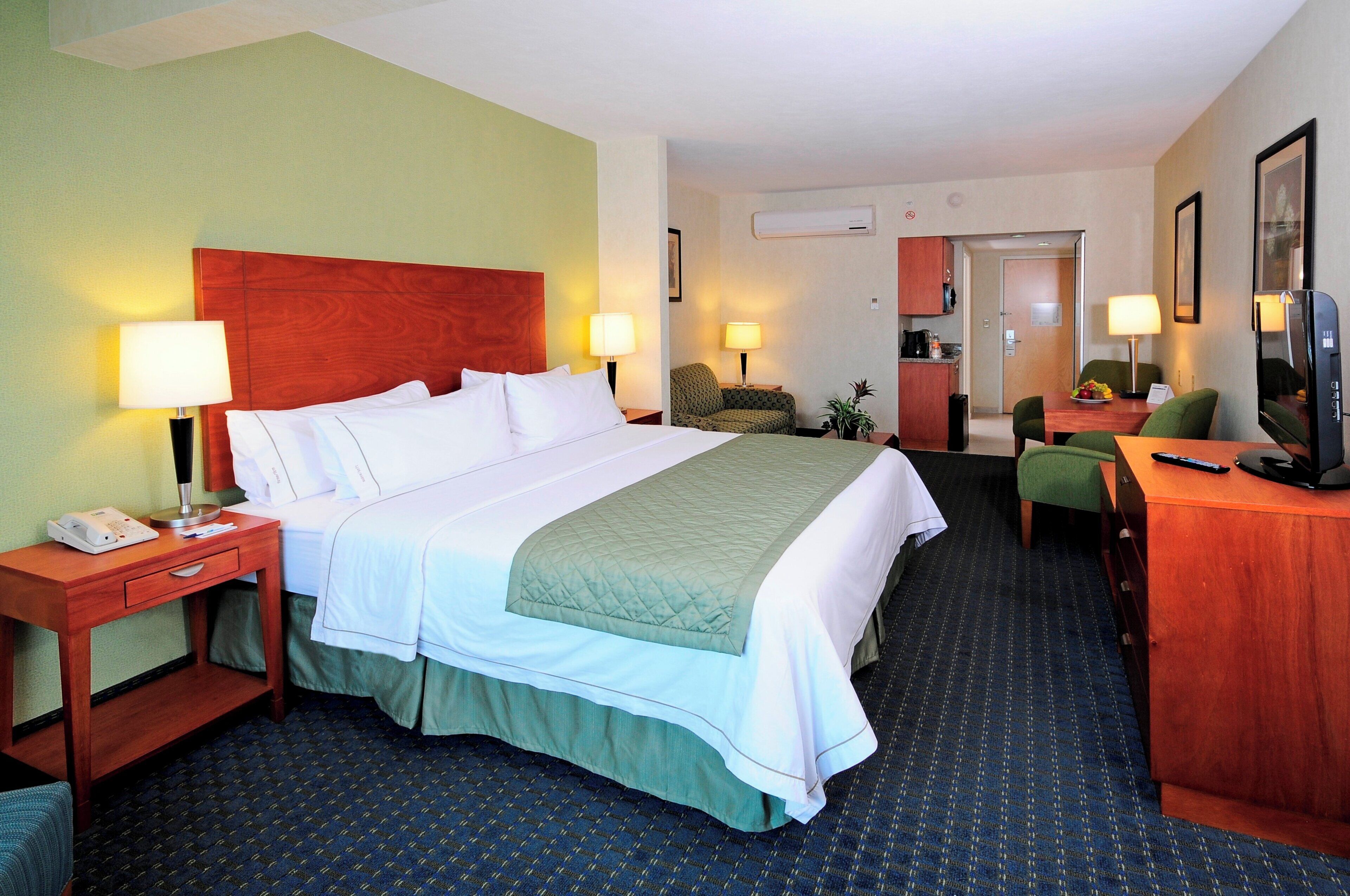 Habitación de hotel con cama, mesitas de noche, escritorio, silla, televisor y kitchenette.
