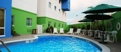 Una piscina con sillas de descanso y sombrillas, rodeada por un edificio de paredes verdes y blancas.
