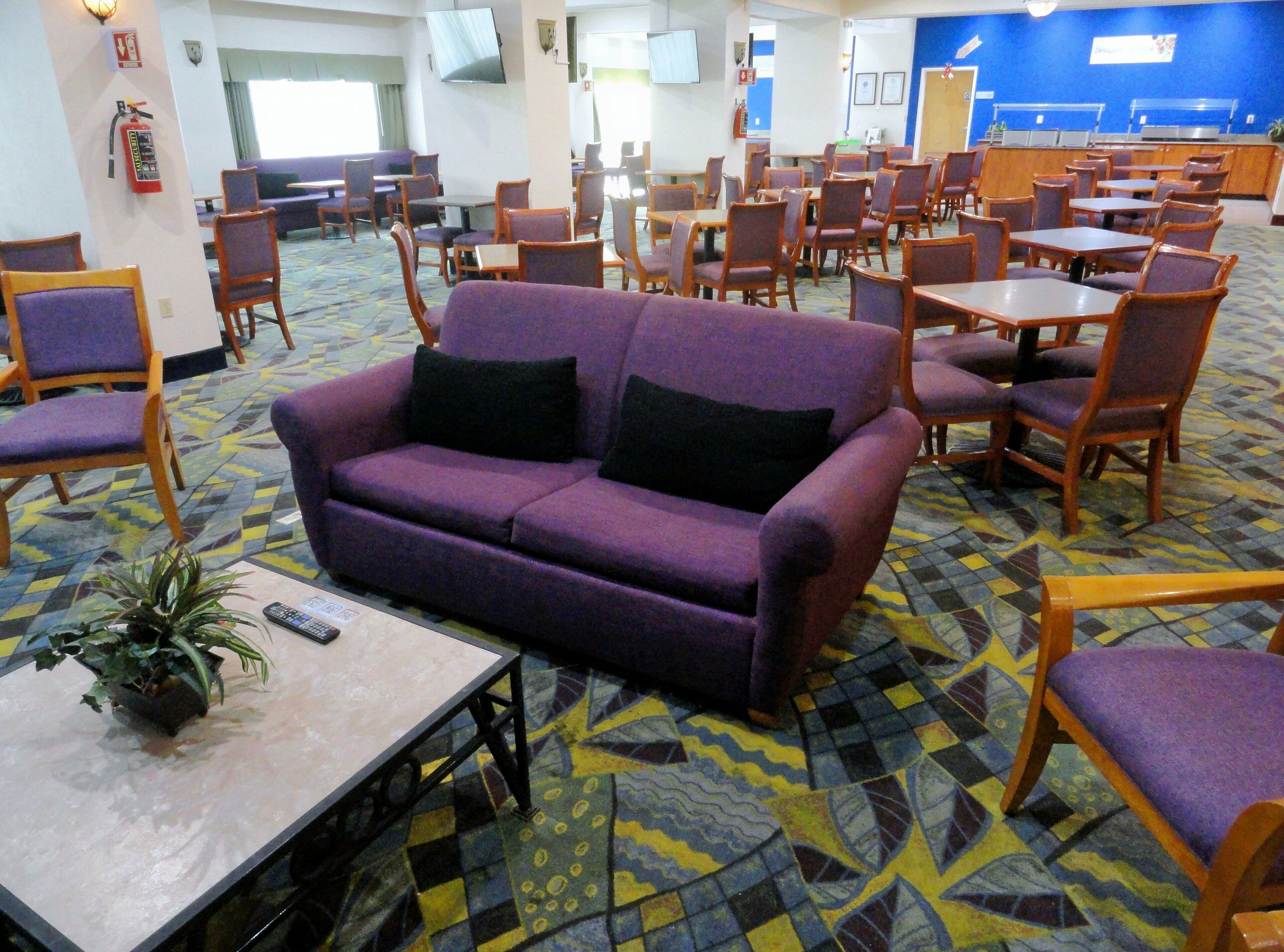 A hotel lobby with purple sofas, wooden tables, and chairs. There is a fire extinguisher on the wall and a television mounted on the wall.