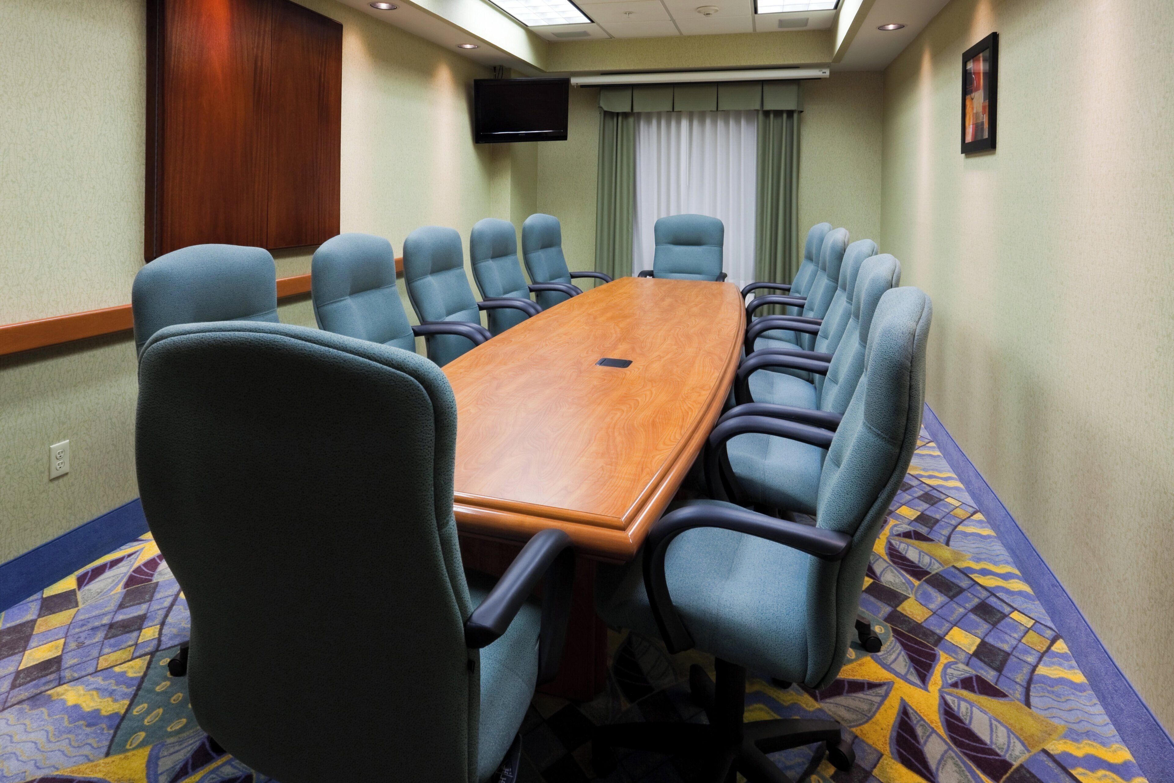 A conference room with a long table and multiple chairs, a flat-screen TV mounted on the wall, and a patterned carpet.