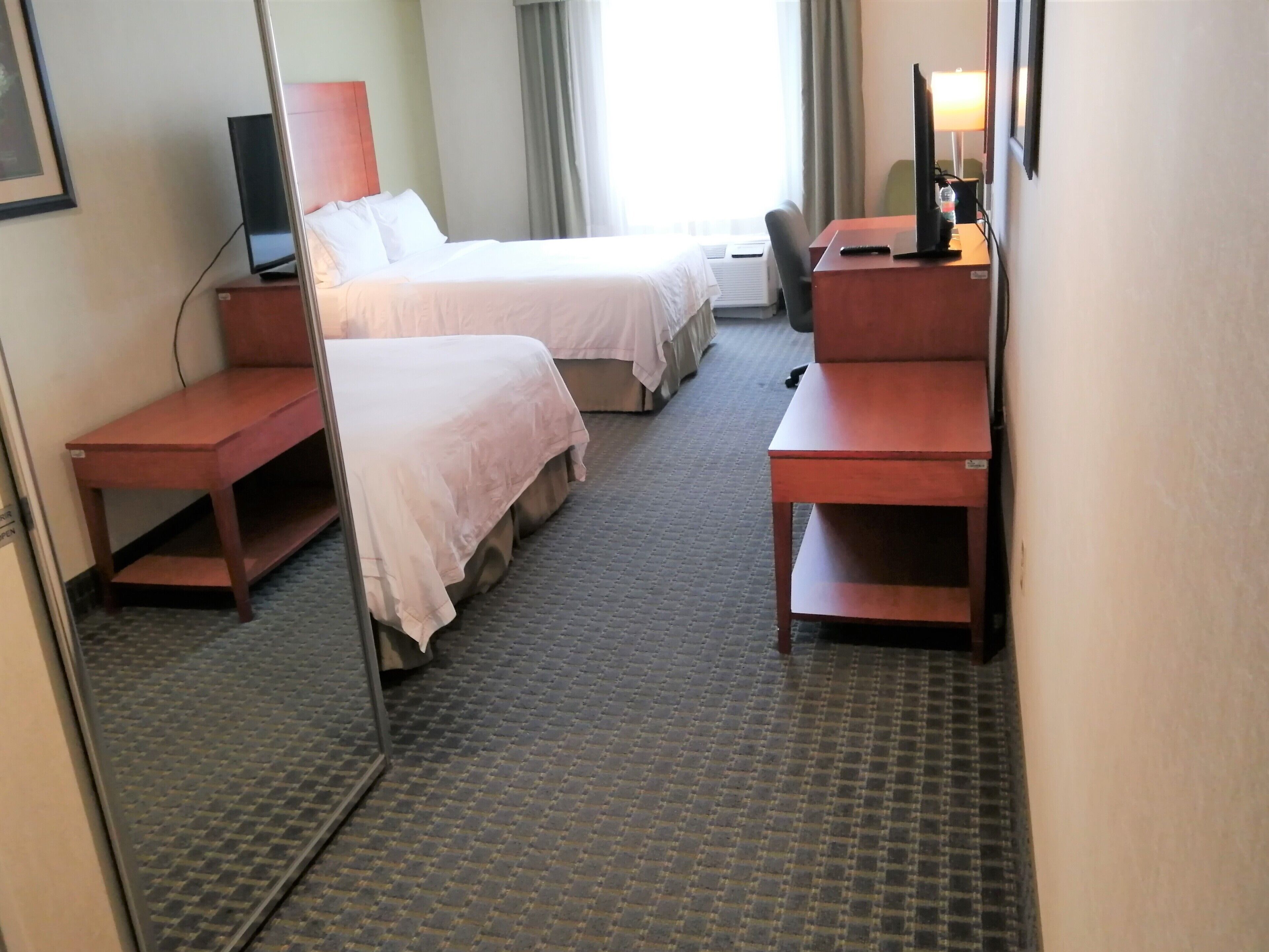 A hotel room with two beds, a desk, a chair, a TV, and a lamp.