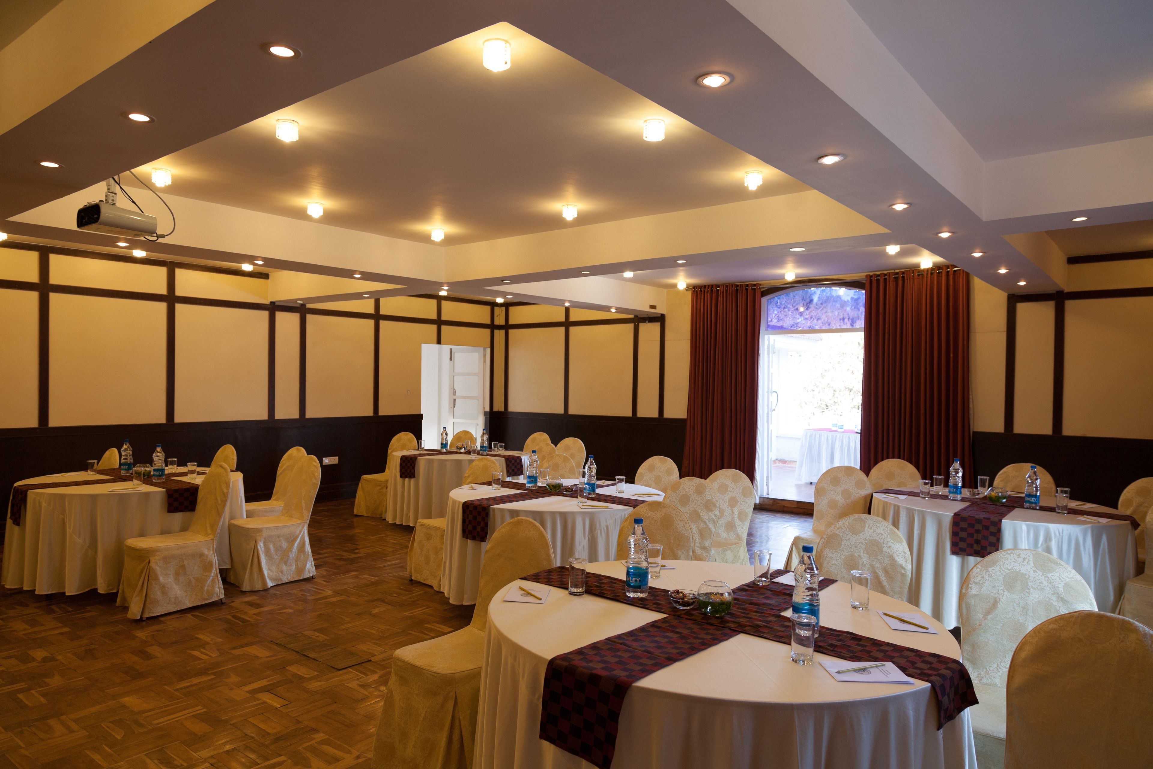 A conference room with multiple round tables, each with white tablecloths and chairs, equipped with water bottles and glasses.