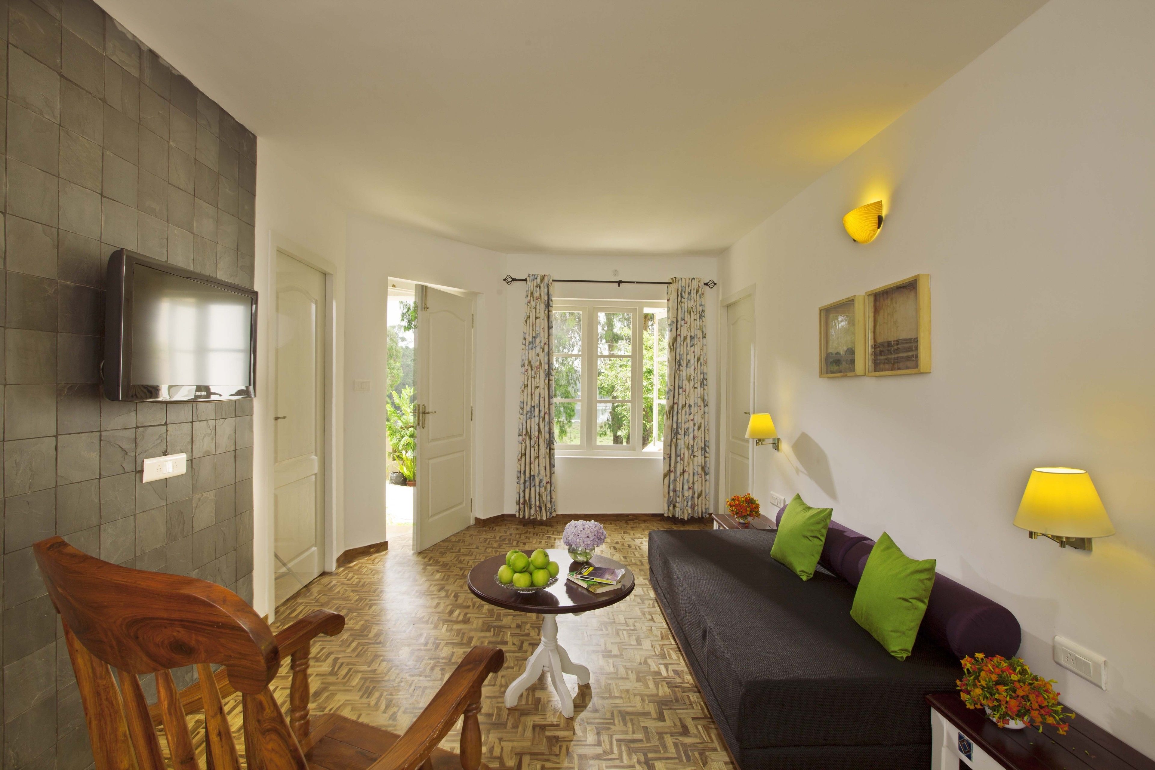 Una sala de estar con un sofá, una silla de madera, una mesa con fruta y un florero, un televisor montado en la pared y una ventana con cortinas.